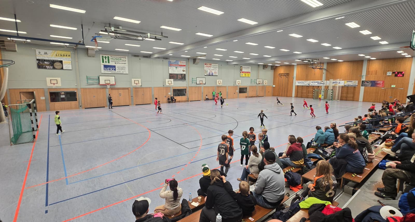 Kinder spielen Hallenfußball; Familien schauen von der Tribüne zu.