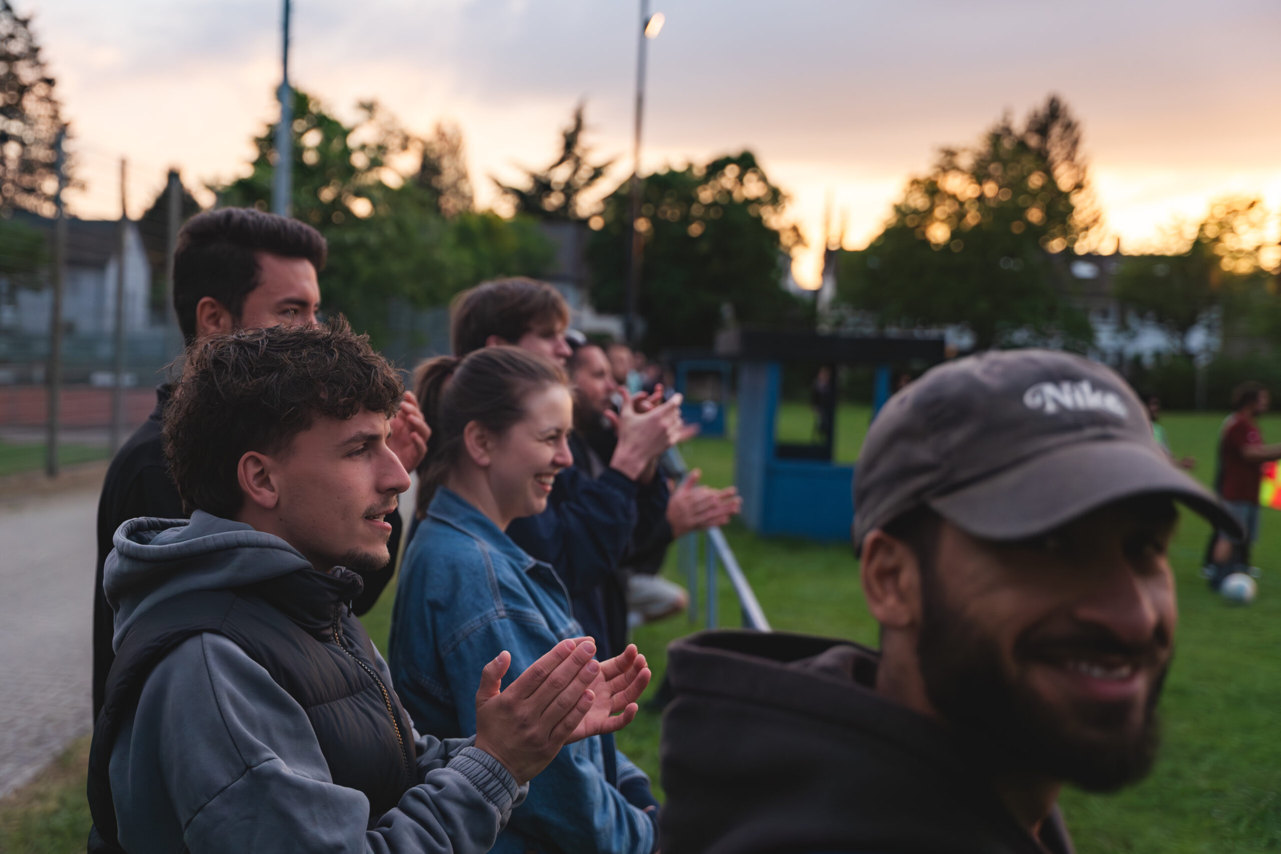 Zuschauer und Mannschaftsmitglieder applaudieren am Spielfeldrand im Abendlicht.